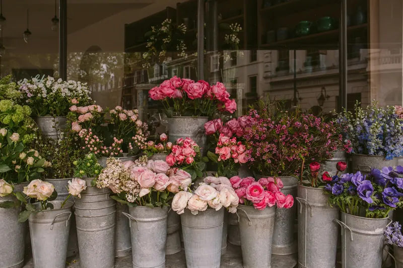Des seaux en zinc remplis de roses roses et blanches et d’anémones violettes, devant la vitrine